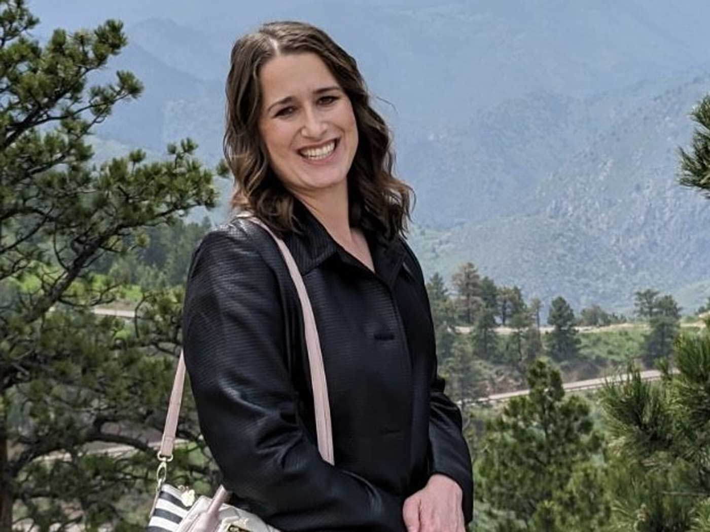 woman with brown hair and wearing a black blazer posing in front of trees and a mountain