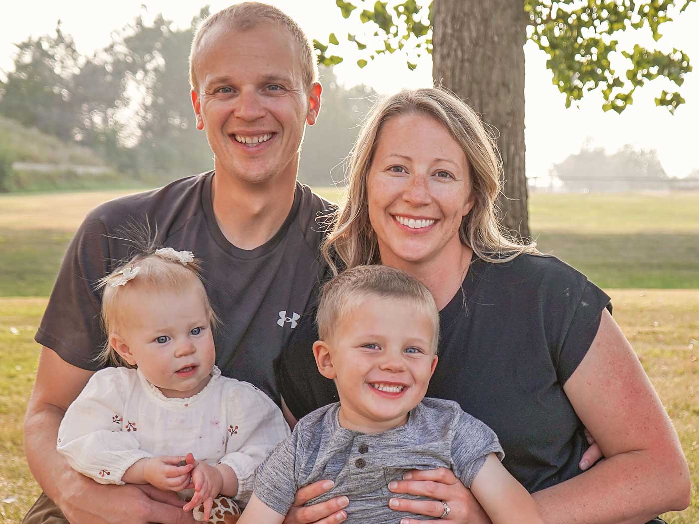 husband and wife with two small children posing outside under a tree
