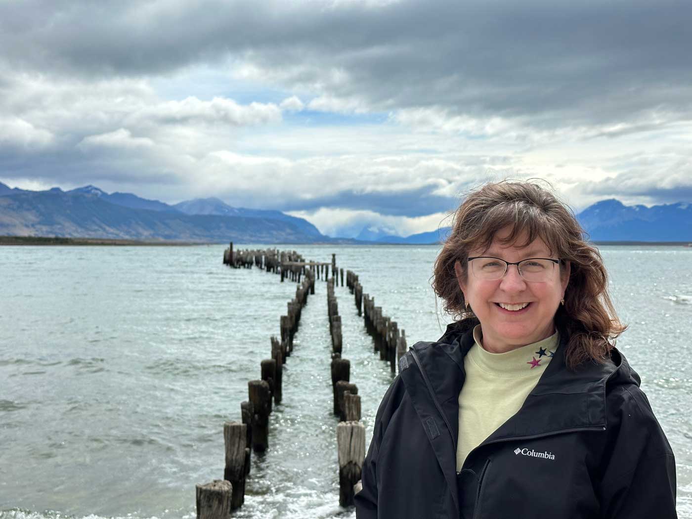 woman wearing black coat standing on pier of lake with mountains in background