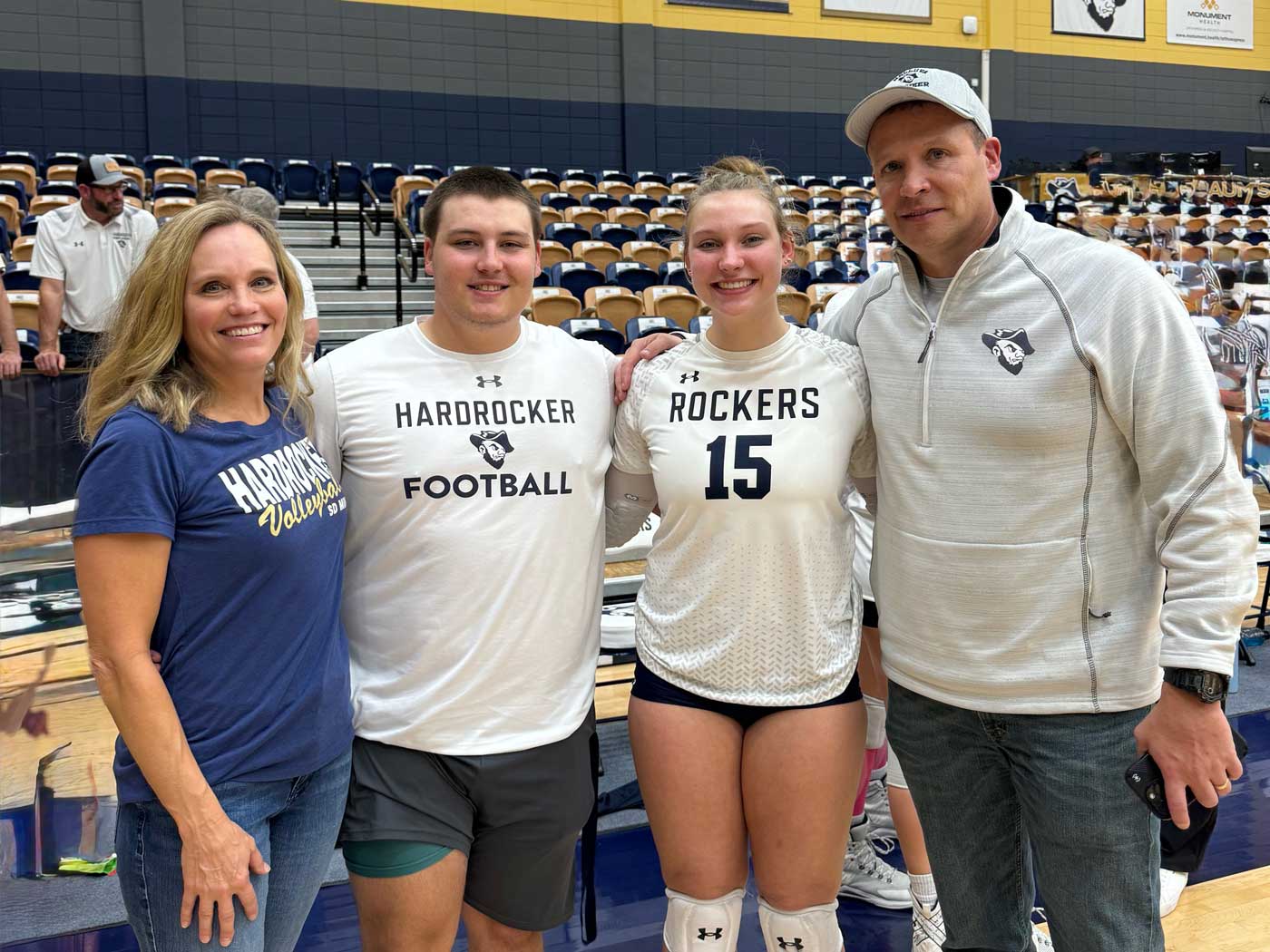 Wickersham-Hardrocker-Family-for-2025-Newsletter-web mother, father, son, and daughter wearing athletic apparel standing on volleyball court with bleachers in background