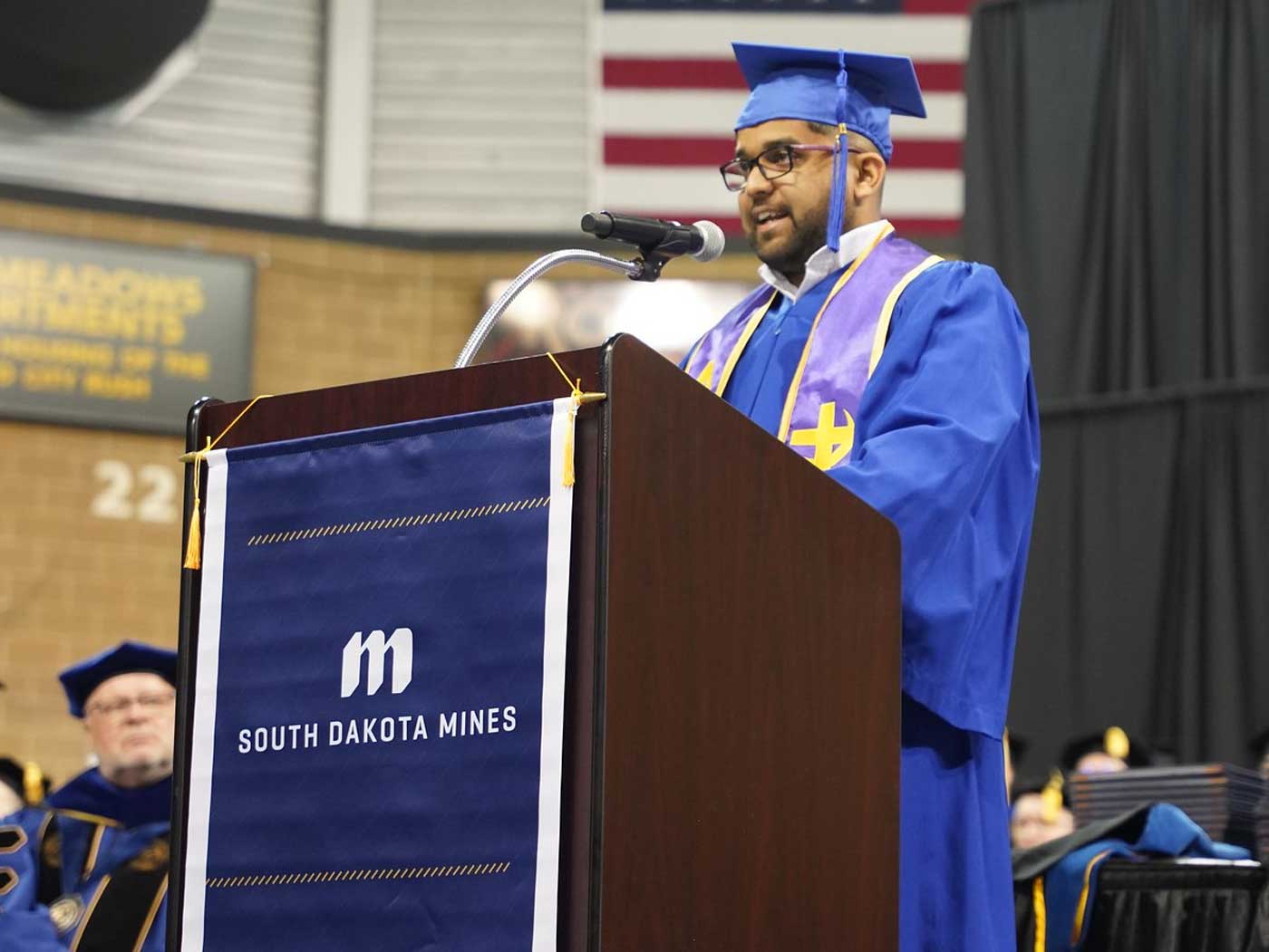 indian man wearing blue graduation gown and cap speaking at a podium