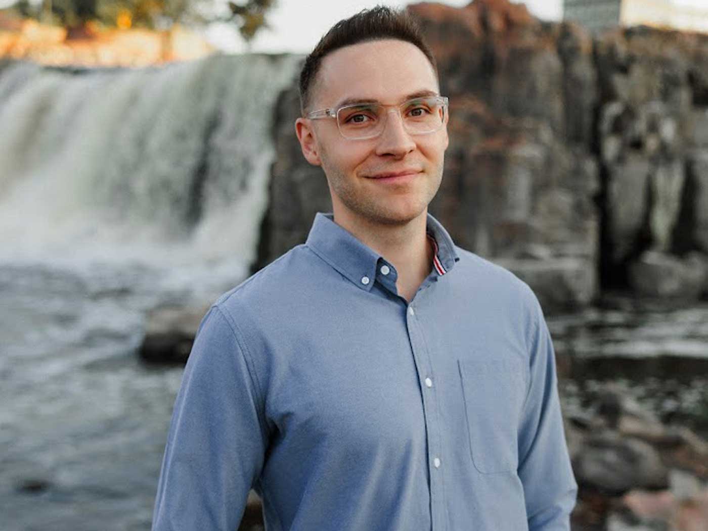 jeremy-feist-web headshot of man wearing glasses and a blue button up shirt in front of a waterfall