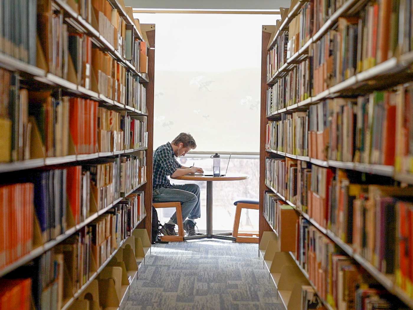 a student at the end of an aisle of books in a library sitting at a table studying