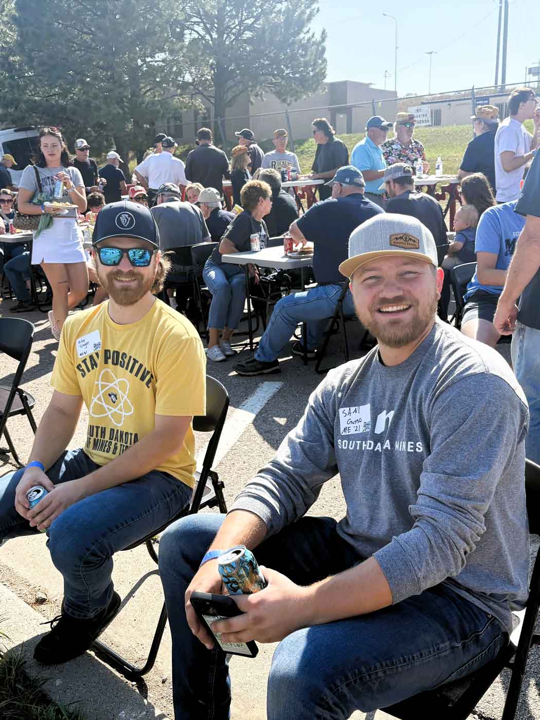 two young men sitting outside in folding chairs holding drinks at a tailgate event